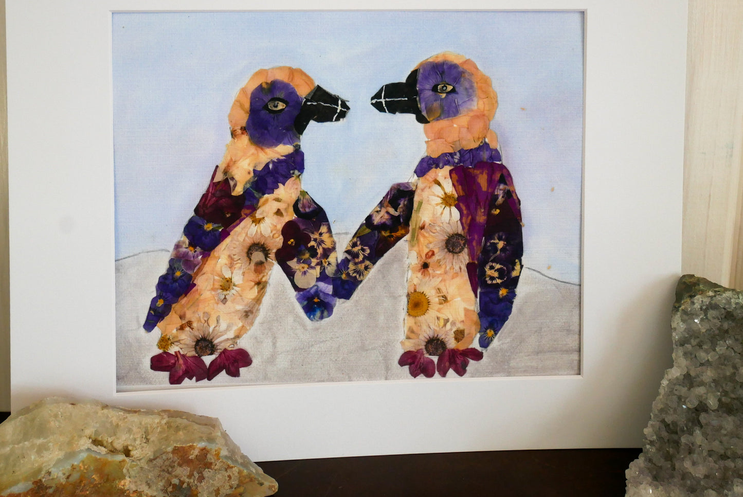 The image is a framed artwork depicting two colorful birds, possibly penguins, with flower petals on their bodies. The background of the artwork is a pale blue color.