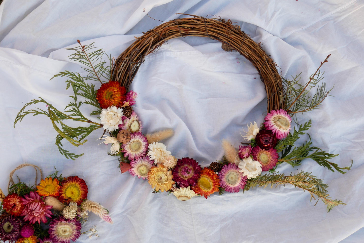 A wreath made of dried flowers and greenery is displayed on a white cloth.