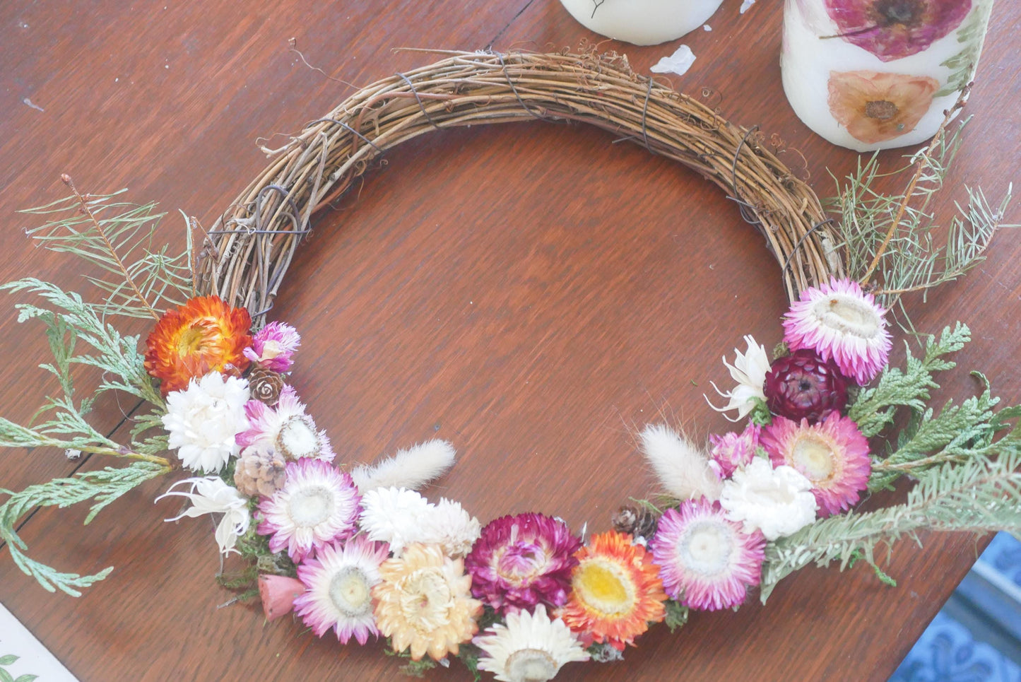 A wreath made of dried flowers and greenery is displayed on a wooden surface, with a white candle and a green plant in the background.