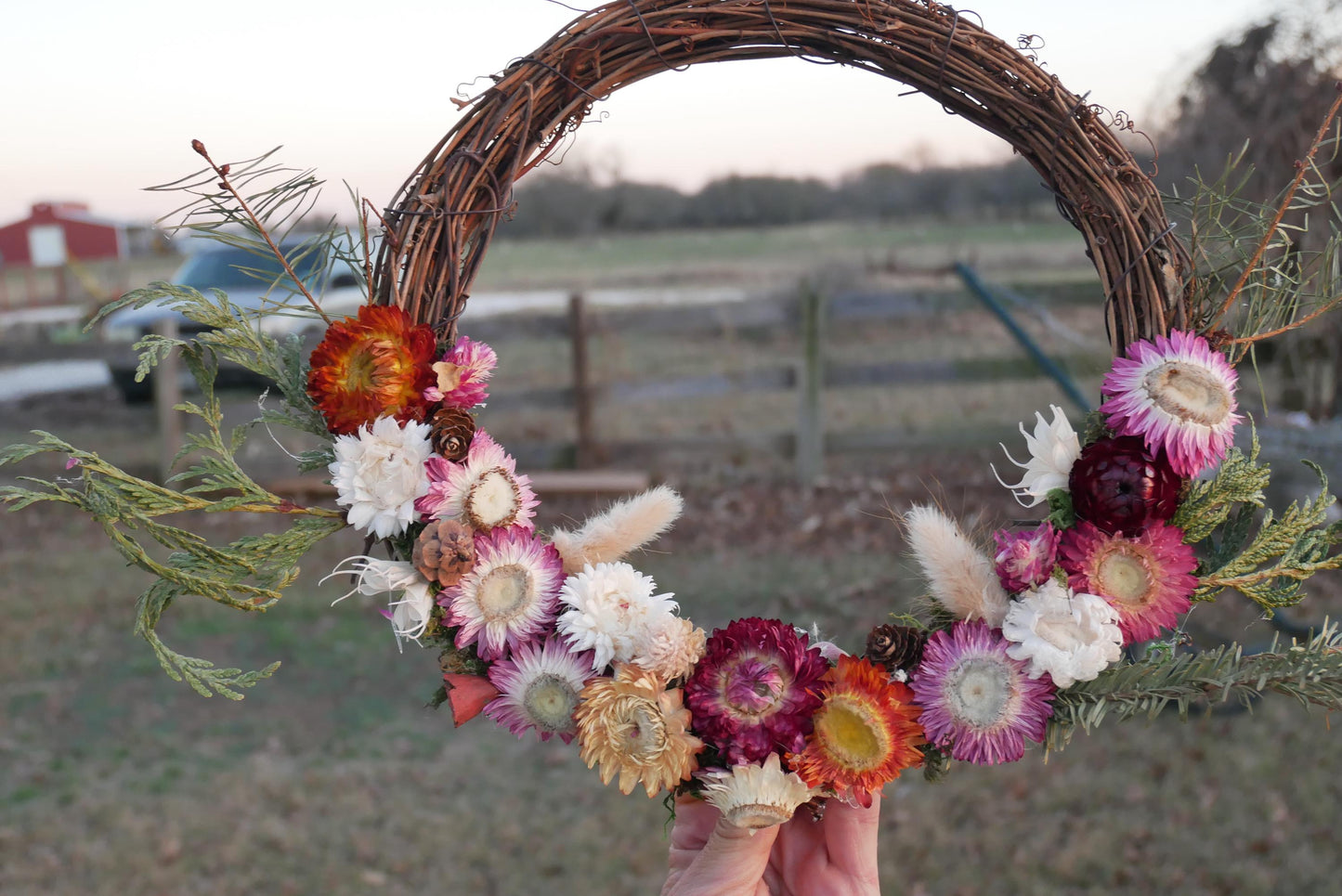 A person holds a wreath made of various colorful flowers, including red, white, and purple blooms, against a backdrop of a rural landscape with a fence and trees.