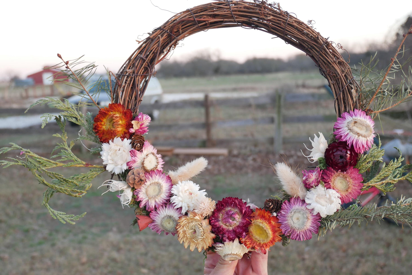 A hand holds a wreath made of dried flowers and branches, with a rustic wooden fence and a red building visible in the background.