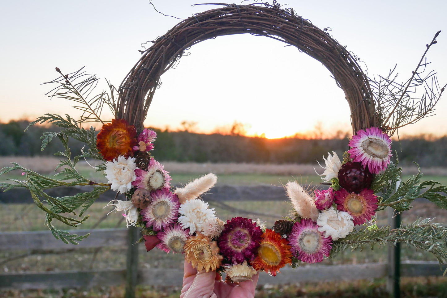 A hand holds a wreath made of dried flowers and greenery, with a field and sunset in the background.
