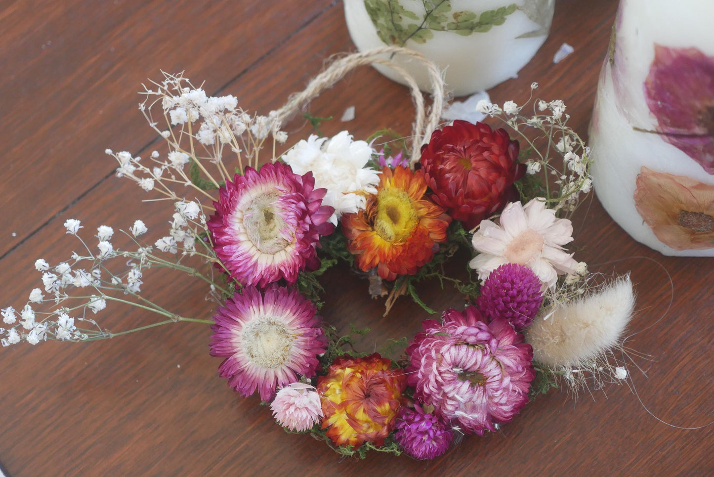 A wreath made of dried flowers, including various colors such as pink, red, white, and purple, is displayed on a wooden surface.