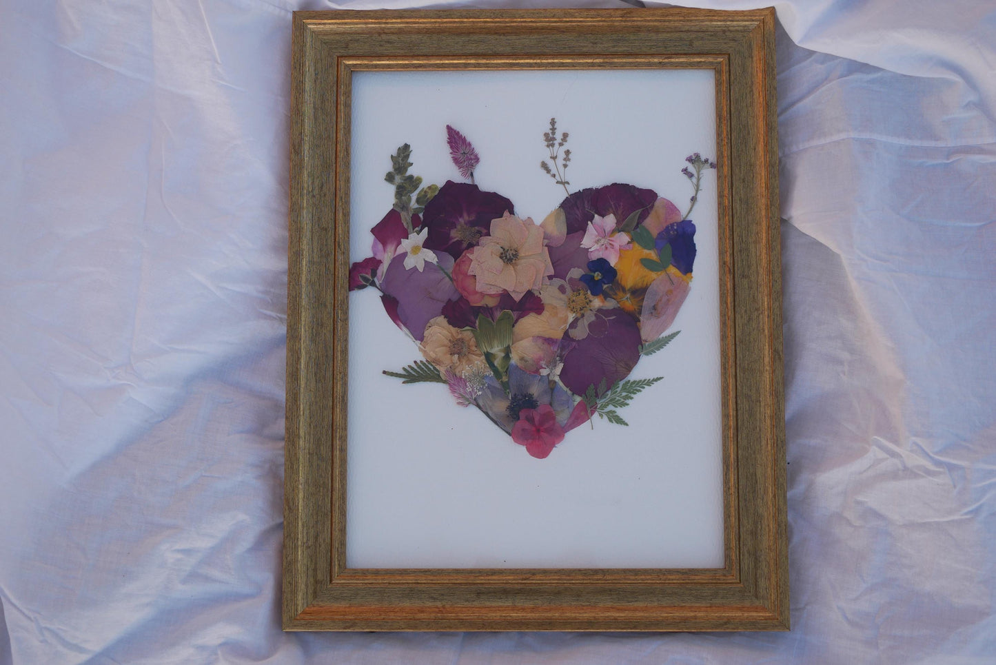 A framed image of a heart made of pressed flowers, including various types of flowers and leaves, is displayed on a white background.
