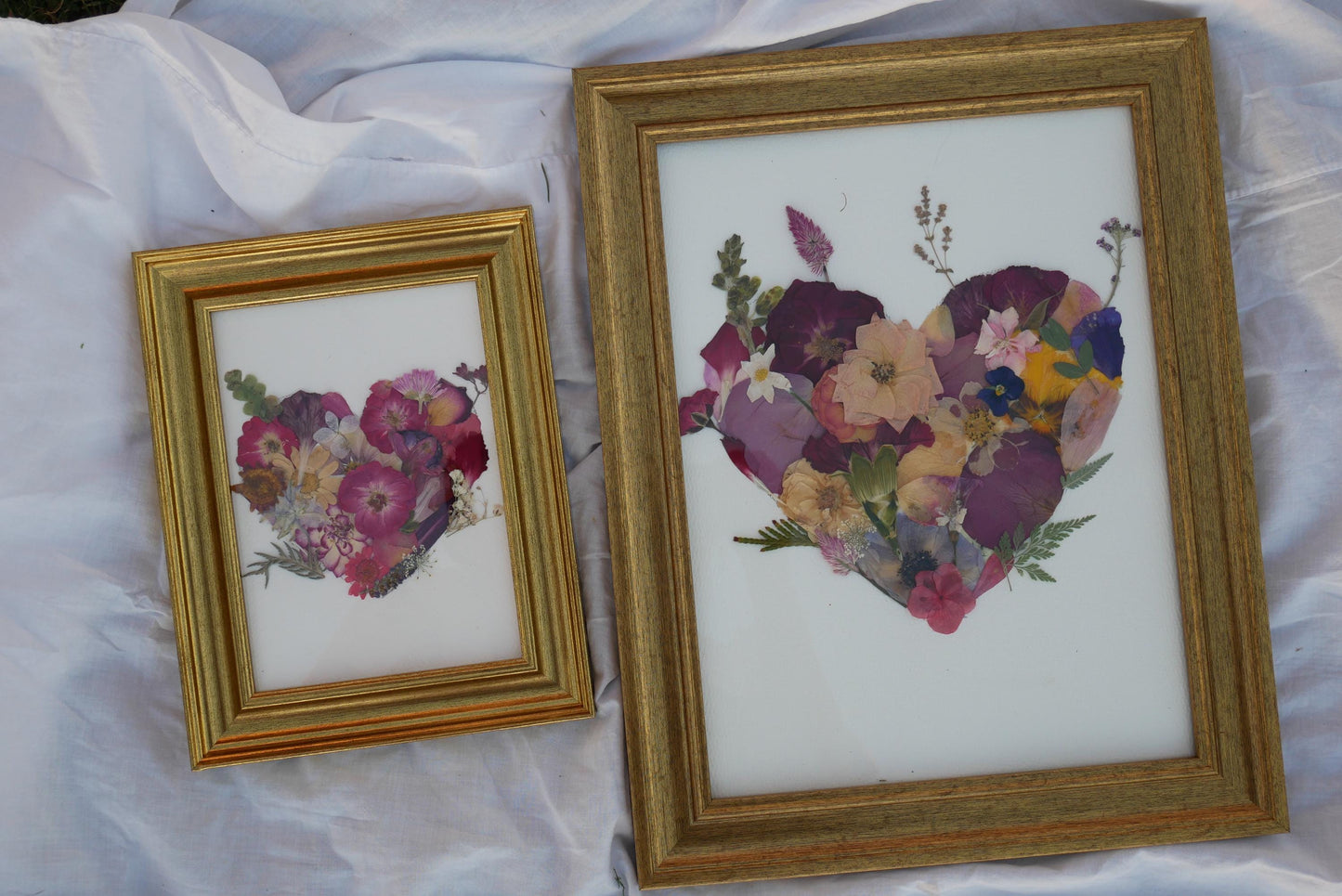 Two framed artworks featuring a heart-shaped arrangement of flowers are displayed on a white cloth.