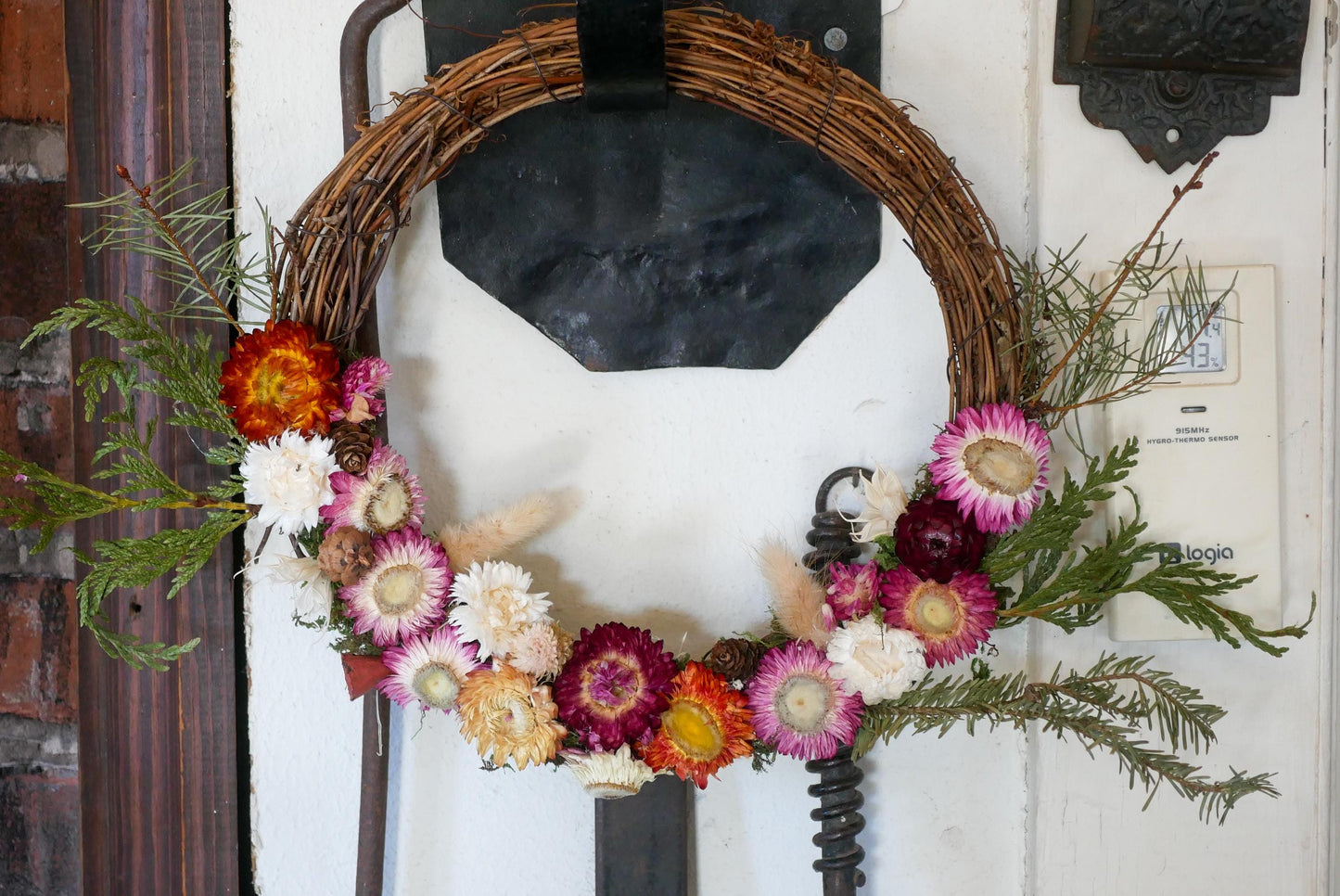 A wreath made of dried flowers and greenery hangs on a wall, with a black metal bracket attached to the wall.