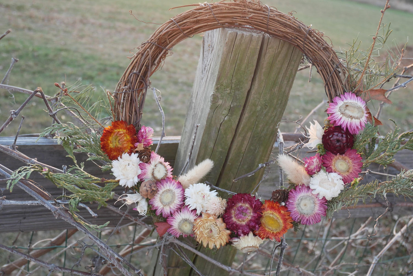 A wreath made of dried flowers and branches is hanging on a wooden fence post.
