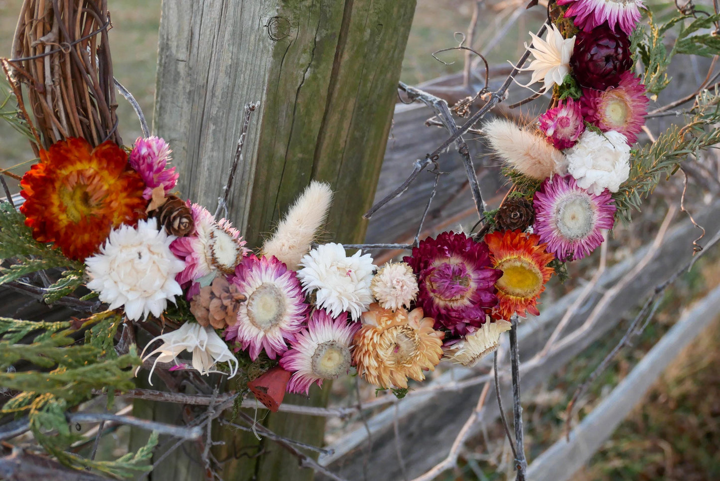 A wreath made of dried flowers, including various colors such as red, white, and orange, is hanging on a wooden fence.