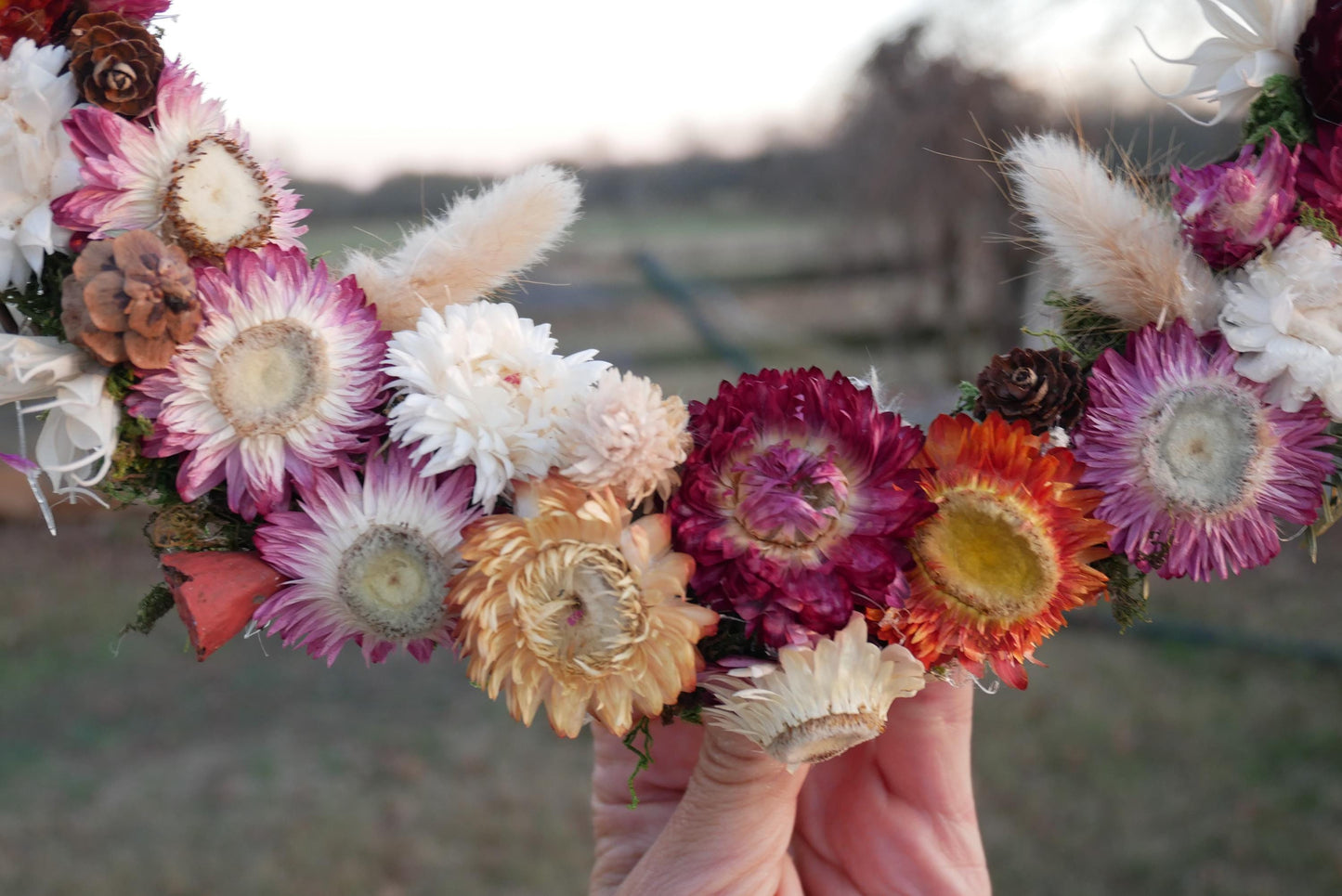 A hand holds a wreath made of dried flowers, including various colors such as pink, white, orange, and purple.