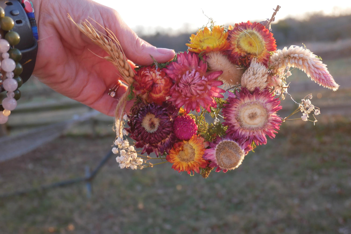 A hand holding a vibrant bouquet of dried flowers, including various types of blooms in shades of pink, yellow, and orange, against a blurred natural background.