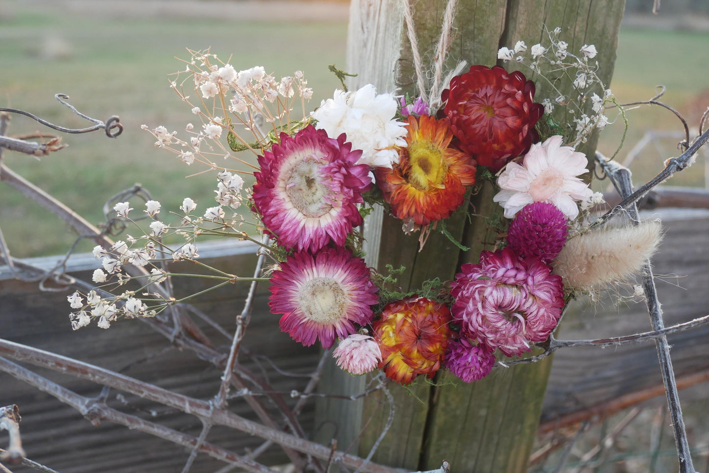 A vibrant bouquet of colorful flowers, including red, orange, and pink blooms, is displayed on a wooden post against a blurred natural background.