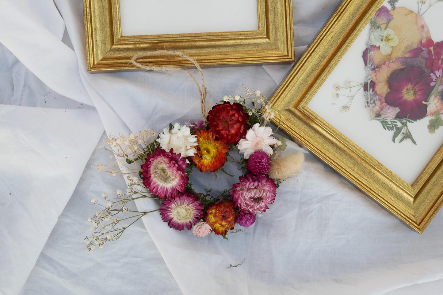 a wreath made of dried flowers and twigs, placed on a white cloth. There are two framed pictures in the background, one with a floral design and the other with a floral pattern.