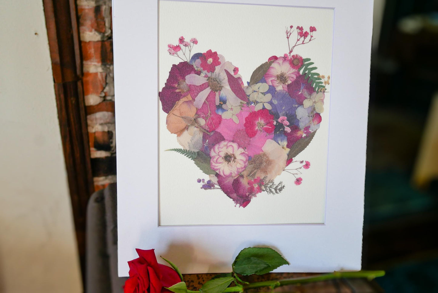 a heart-shaped arrangement of colorful flowers and leaves, displayed in a white frame.