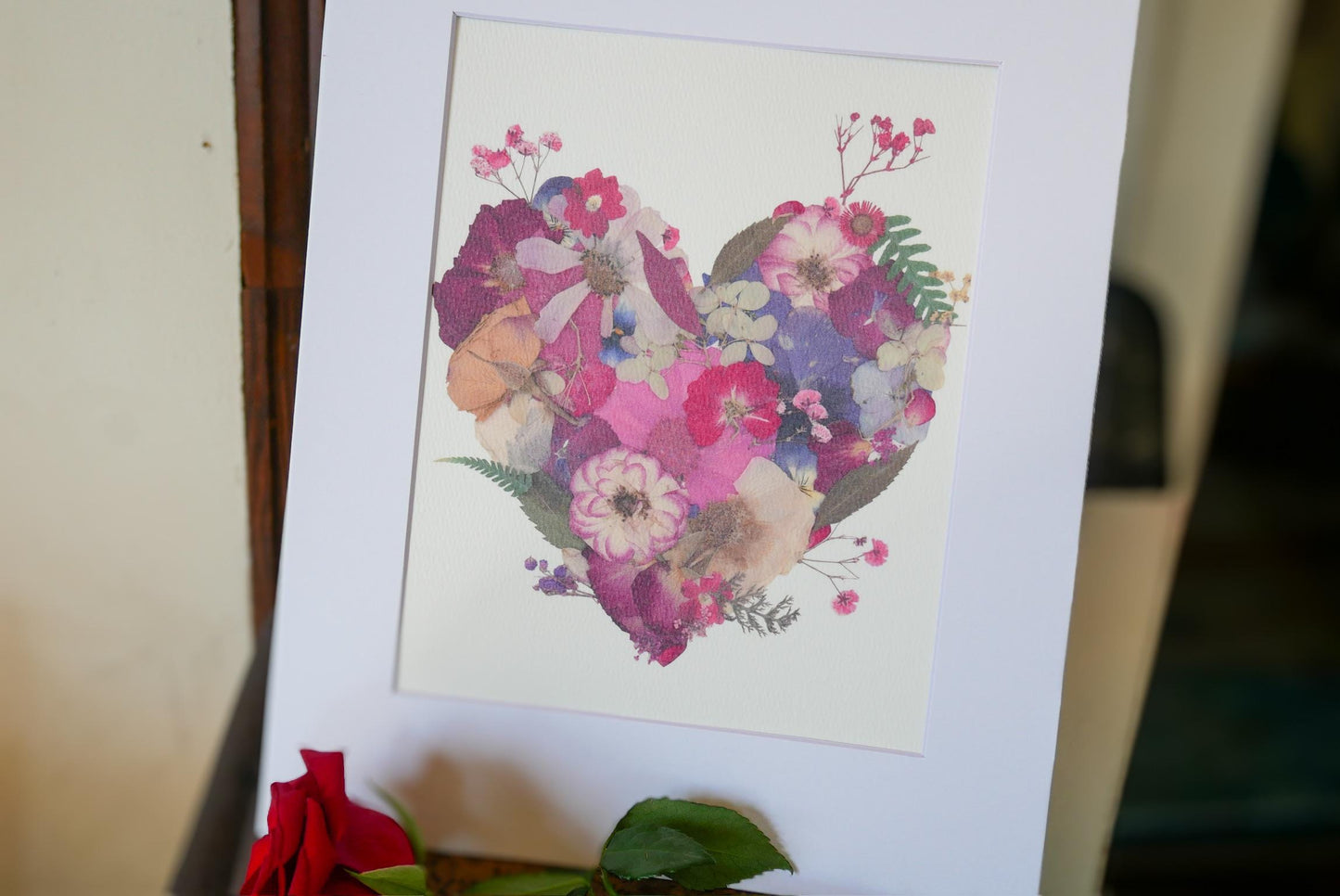 A framed print of a heart-shaped arrangement of colorful flowers, including roses, is displayed on a white surface.