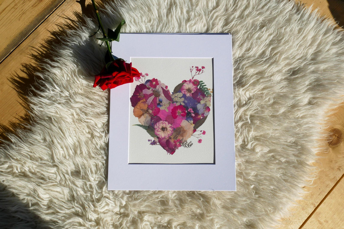 a white frame with a heart-shaped floral print on it, placed on a white furry surface. Next to the frame, there is a red rose.