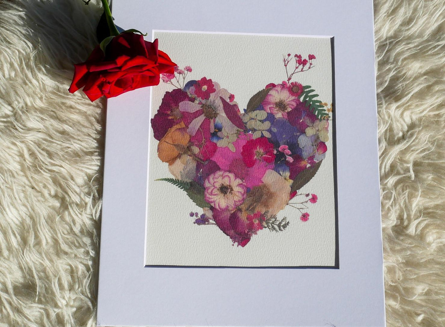 A framed print of a heart-shaped arrangement of colorful flowers is displayed on a white surface, accompanied by a single red rose.