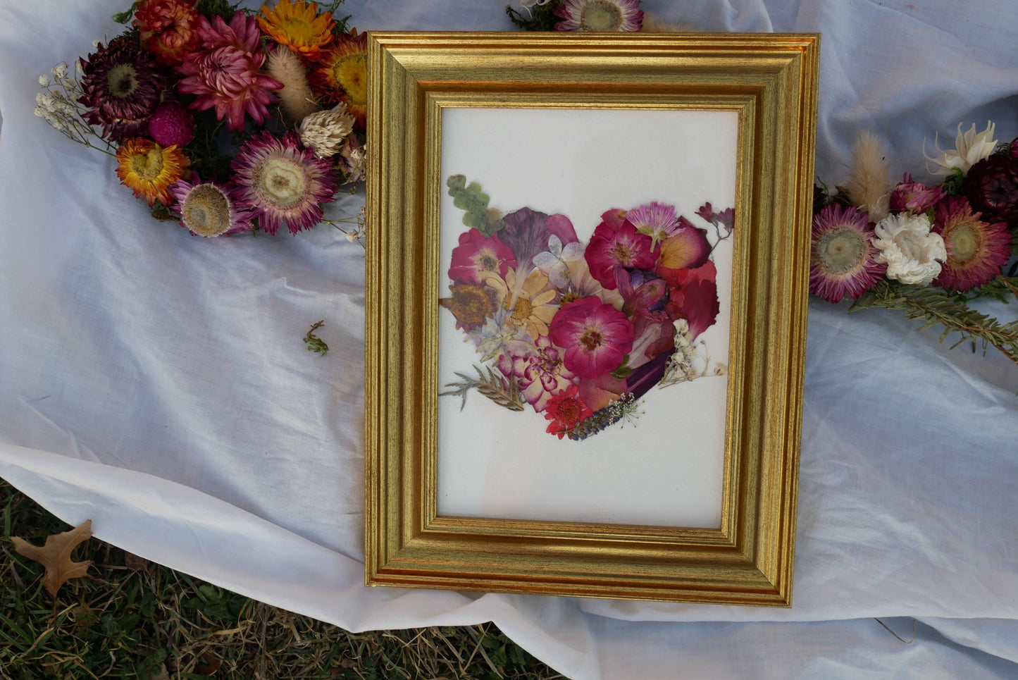 A framed image of a heart-shaped arrangement of dried flowers is displayed on a white cloth, with a bouquet of colorful flowers visible in the background.