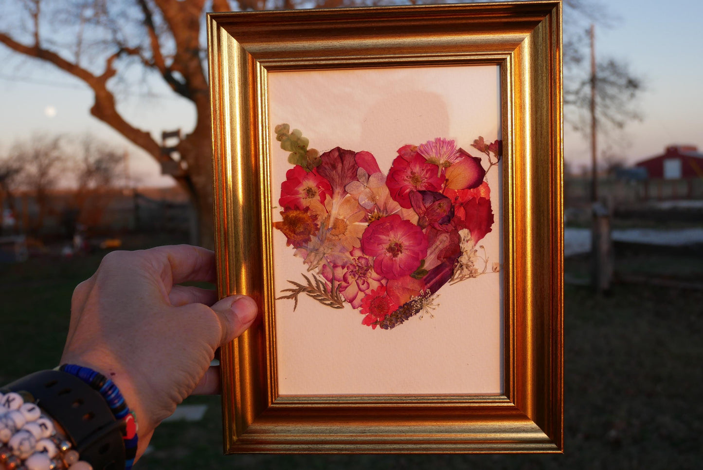 A hand is holding a framed picture of a heart-shaped arrangement of colorful flowers.