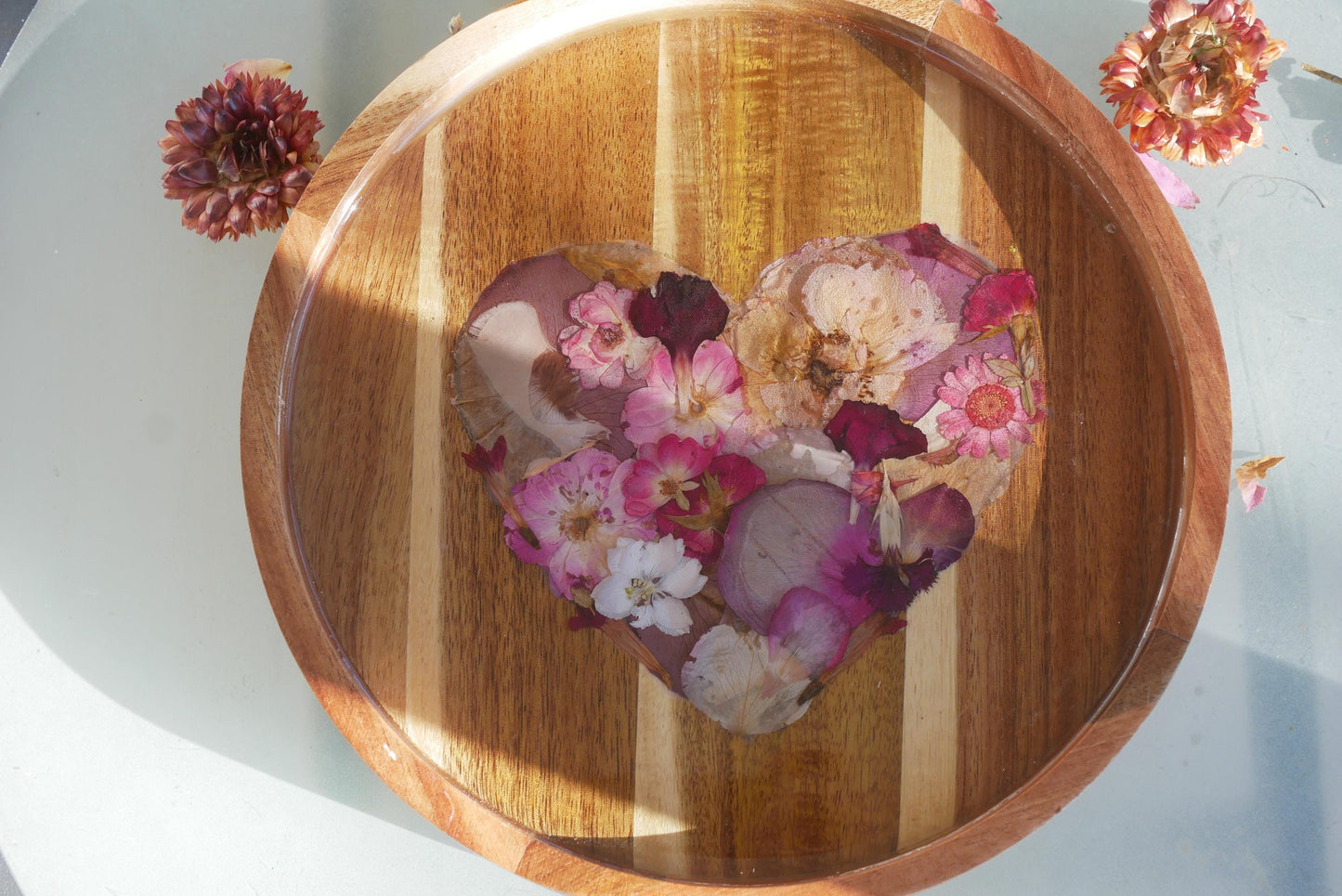 A wooden tray with a heart-shaped arrangement of dried flowers, including pink and white flowers, resting on a white surface.