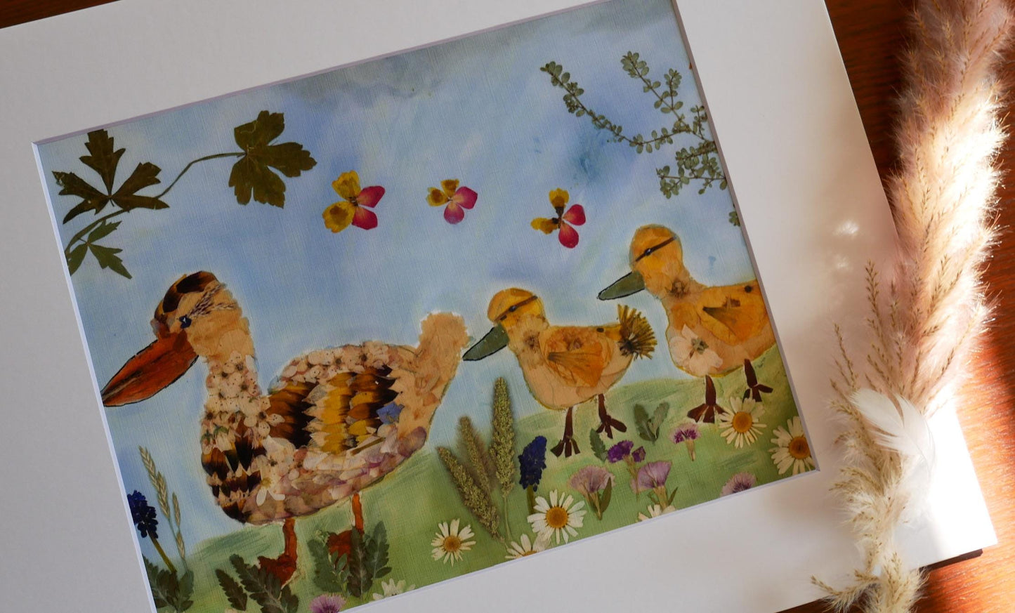 The image is a framed artwork depicting a family of ducks in a field with flowers and plants.