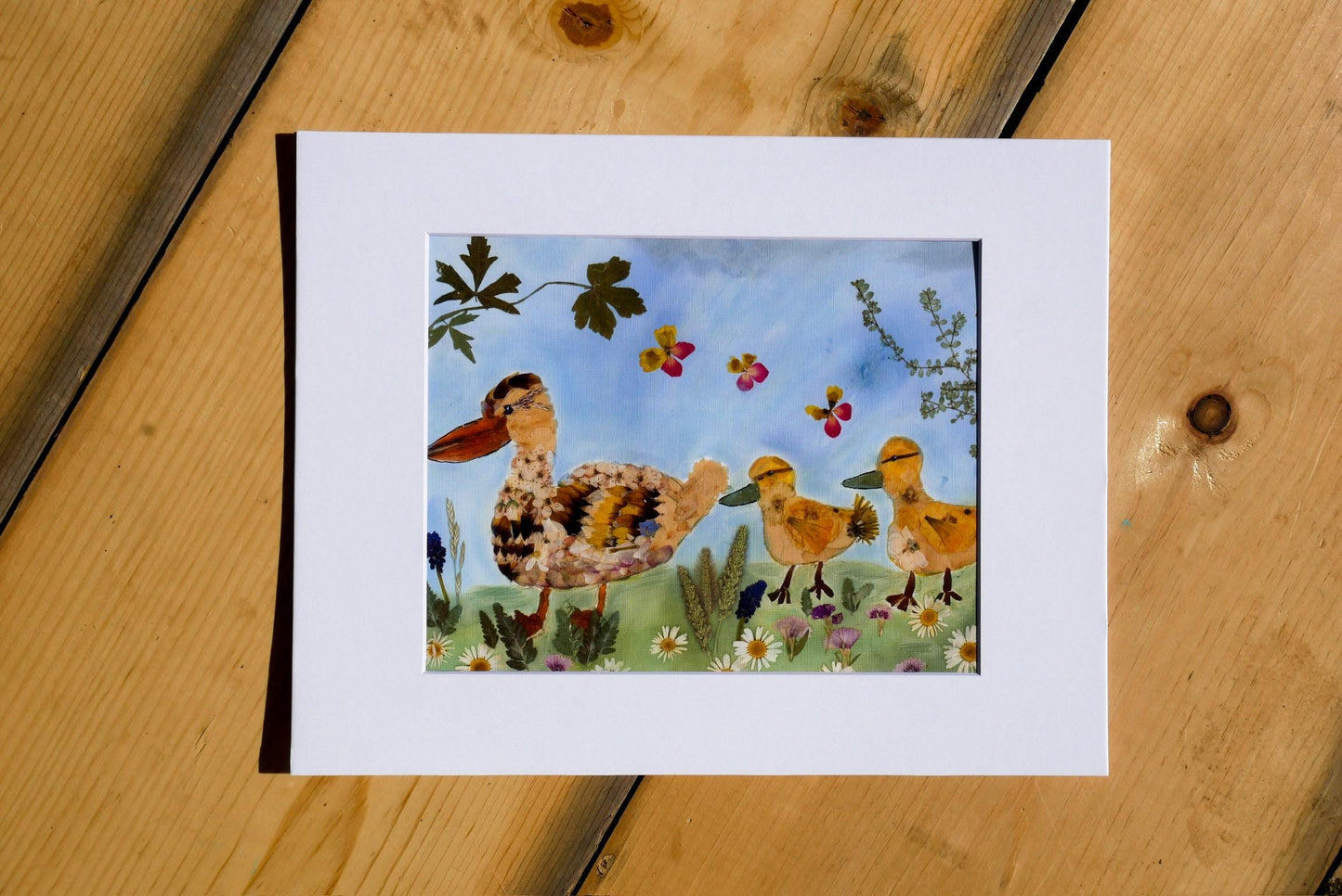 A framed watercolor painting depicting a family of ducks in a grassy field with flowers and leaves.