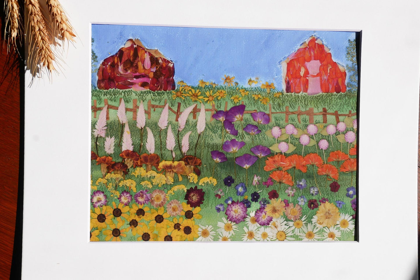The image is a vibrant painting depicting a lush garden filled with a variety of colorful flowers, including sunflowers, daisies, and other blooms. In the background, there are two red barns, adding a rustic charm to the scene. The painting is framed and displayed on a white surface, creating a visually appealing contrast with the colorful flowers and the red barns.