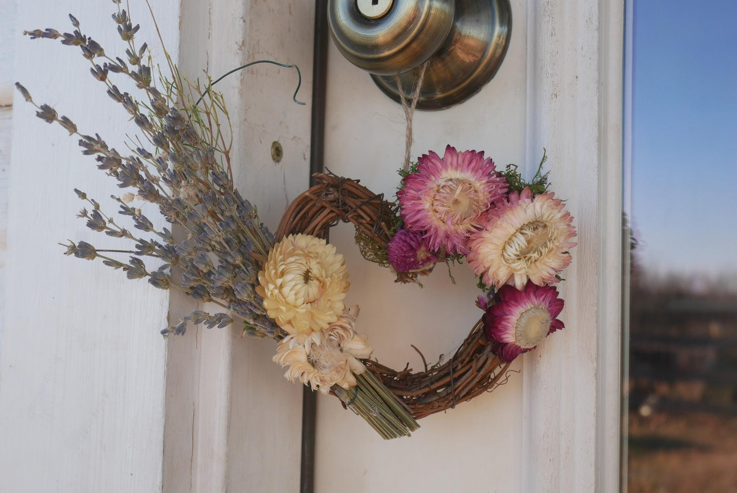 A wreath made of dried flowers and twigs hangs on a white door, with a brass door knob visible above it.