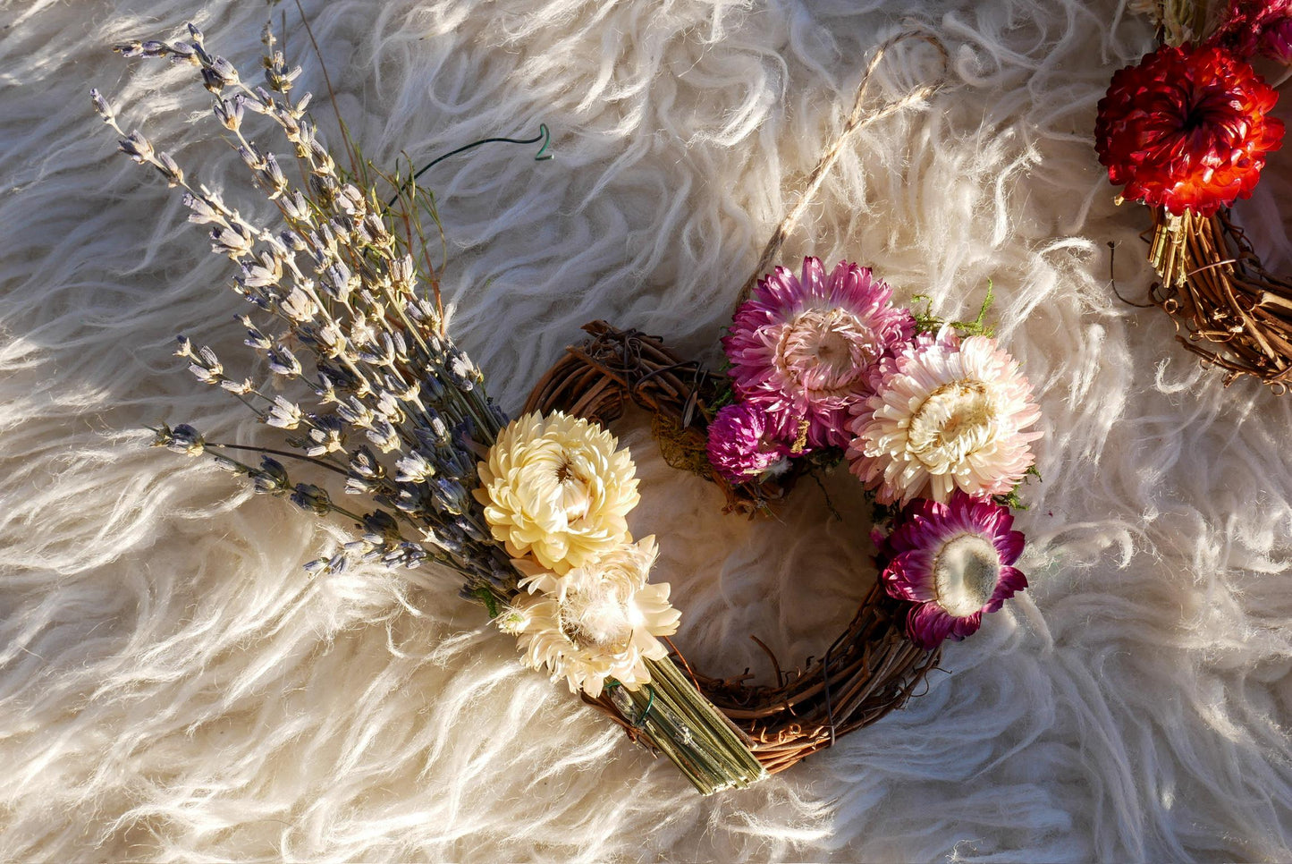 A wreath made of dried flowers, including purple and white flowers, is displayed on a white furry surface.