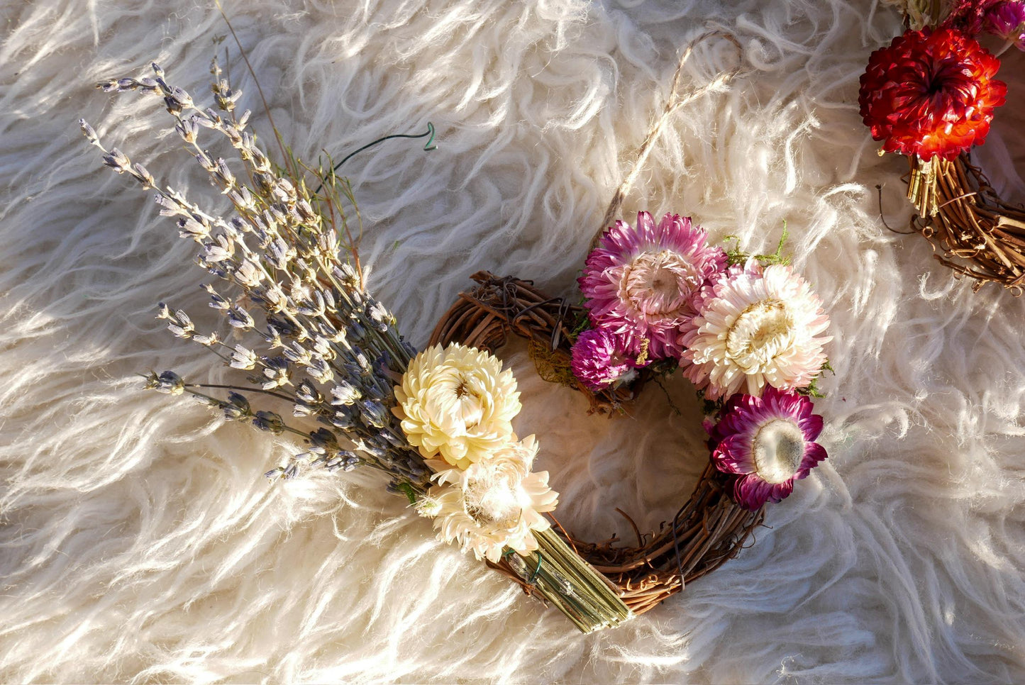 A wreath made of dried flowers and twigs, with a mix of white, purple, and red flowers, resting on a white furry surface.