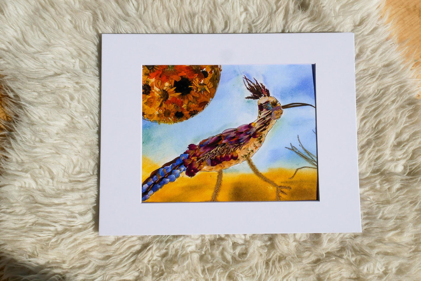 The image is a framed artwork depicting a colorful bird with a long tail, standing on a grassy field with a large sun in the background.