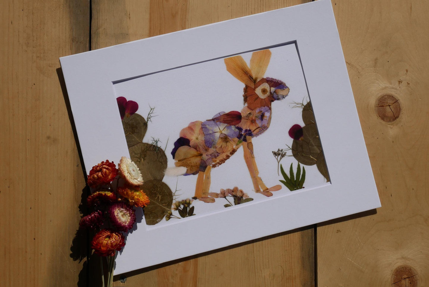 A framed artwork depicting a colorful rabbit surrounded by dried flowers and leaves, placed on a wooden surface.