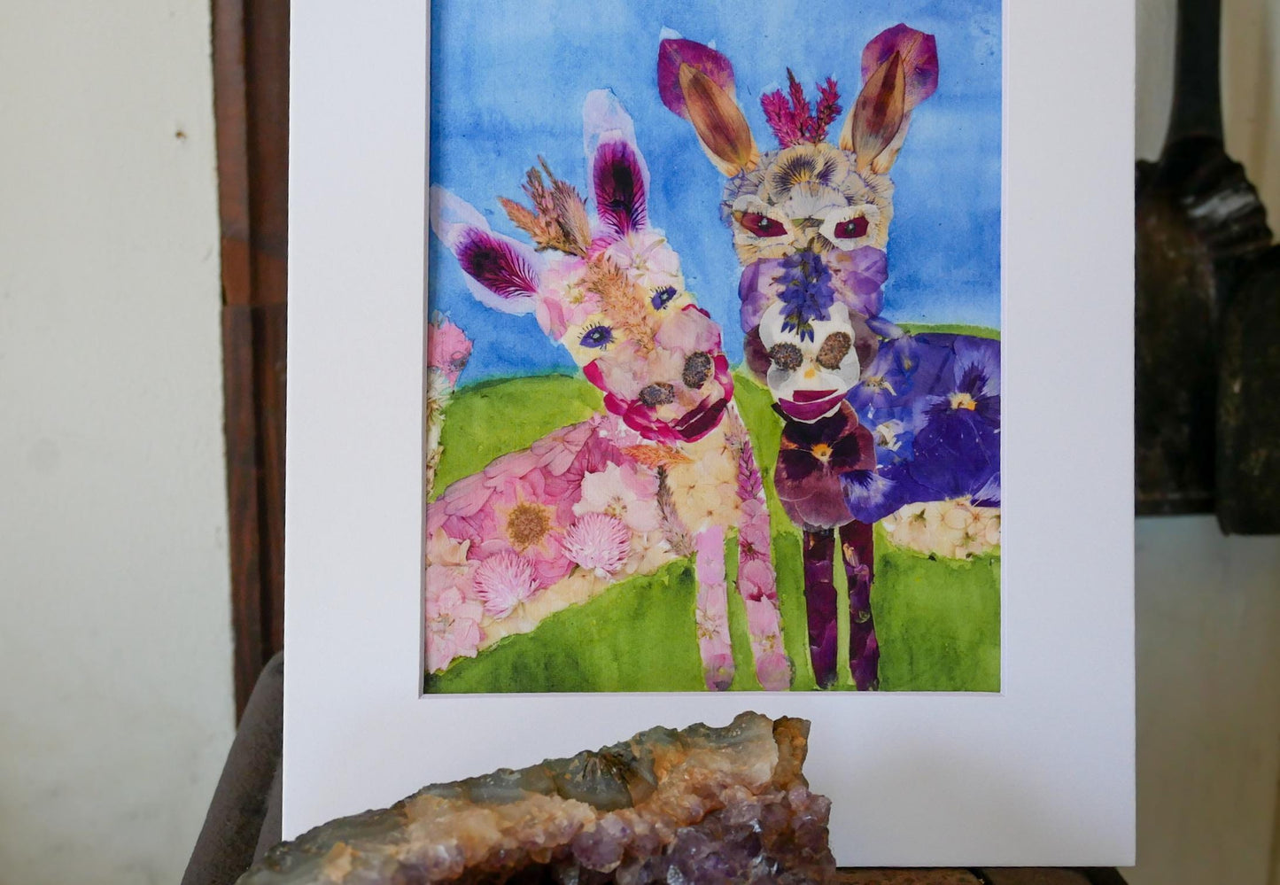 a vibrant painting of two whimsical animals, a donkey and a horse, set against a lush green field. The painting is framed in a white frame and displayed on a shelf, with a piece of rock or stone visible in the foreground.