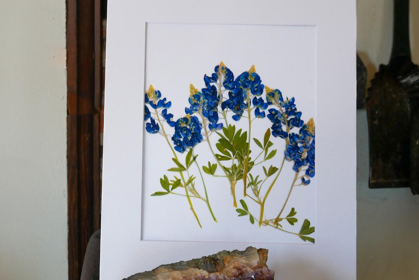 a framed artwork of blue flowers with green leaves, displayed on a white surface.