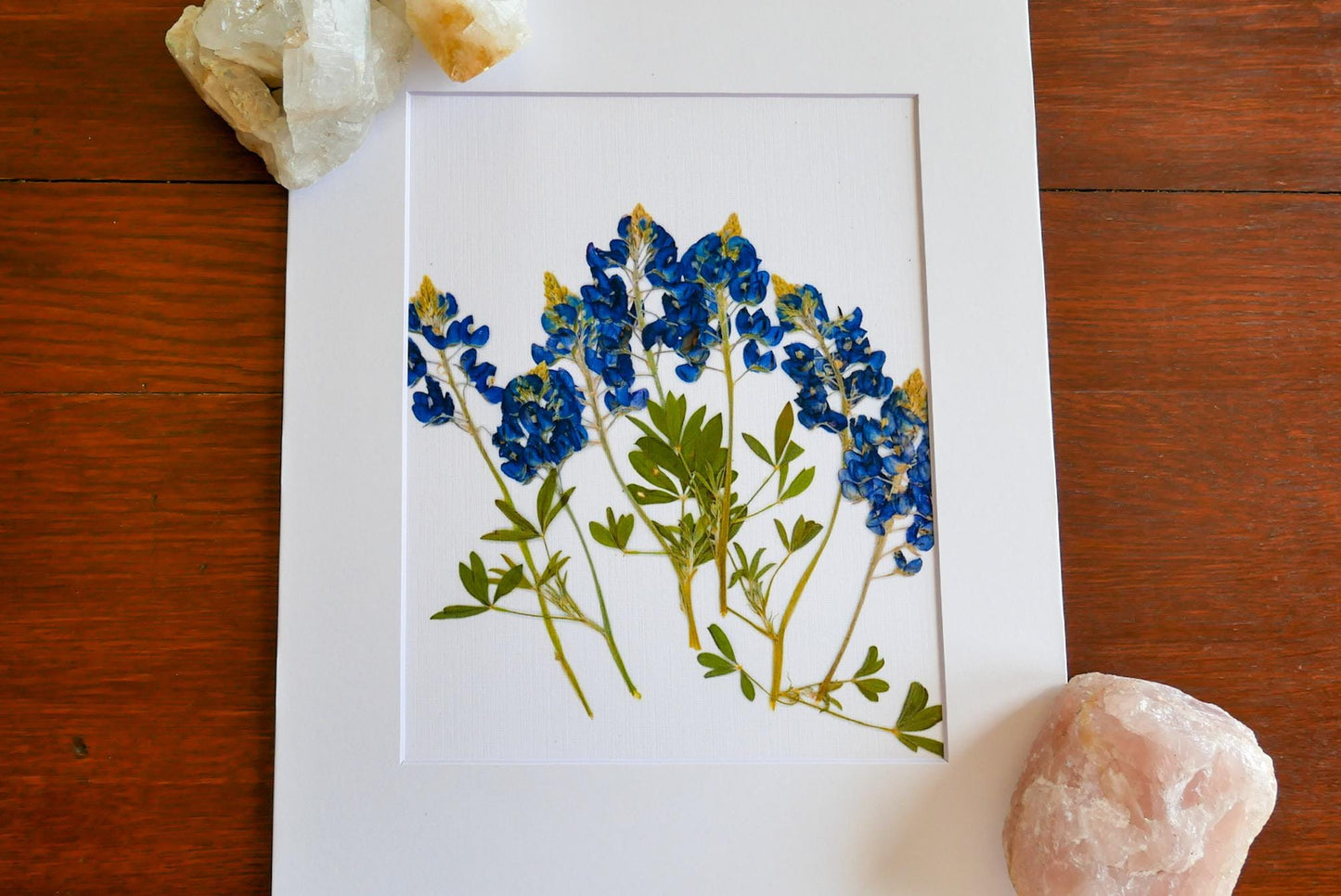 a framed print of blue flowers with green leaves, placed on a wooden surface next to a crystal and a piece of rock.