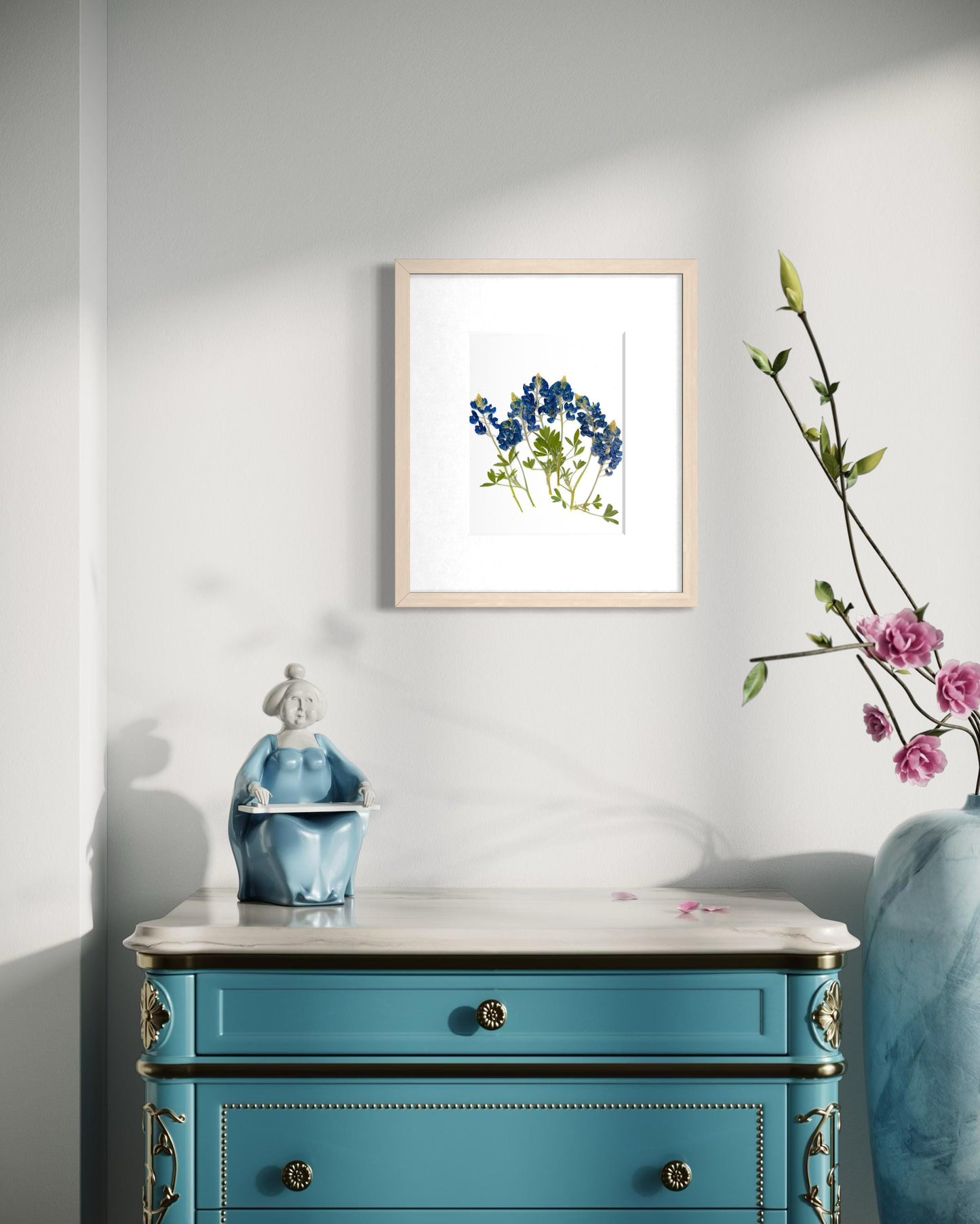 a blue dresser with a framed picture of blue flowers hanging on the wall above it. The dresser has a white marble top and is decorated with a small figurine and a vase of pink flowers.