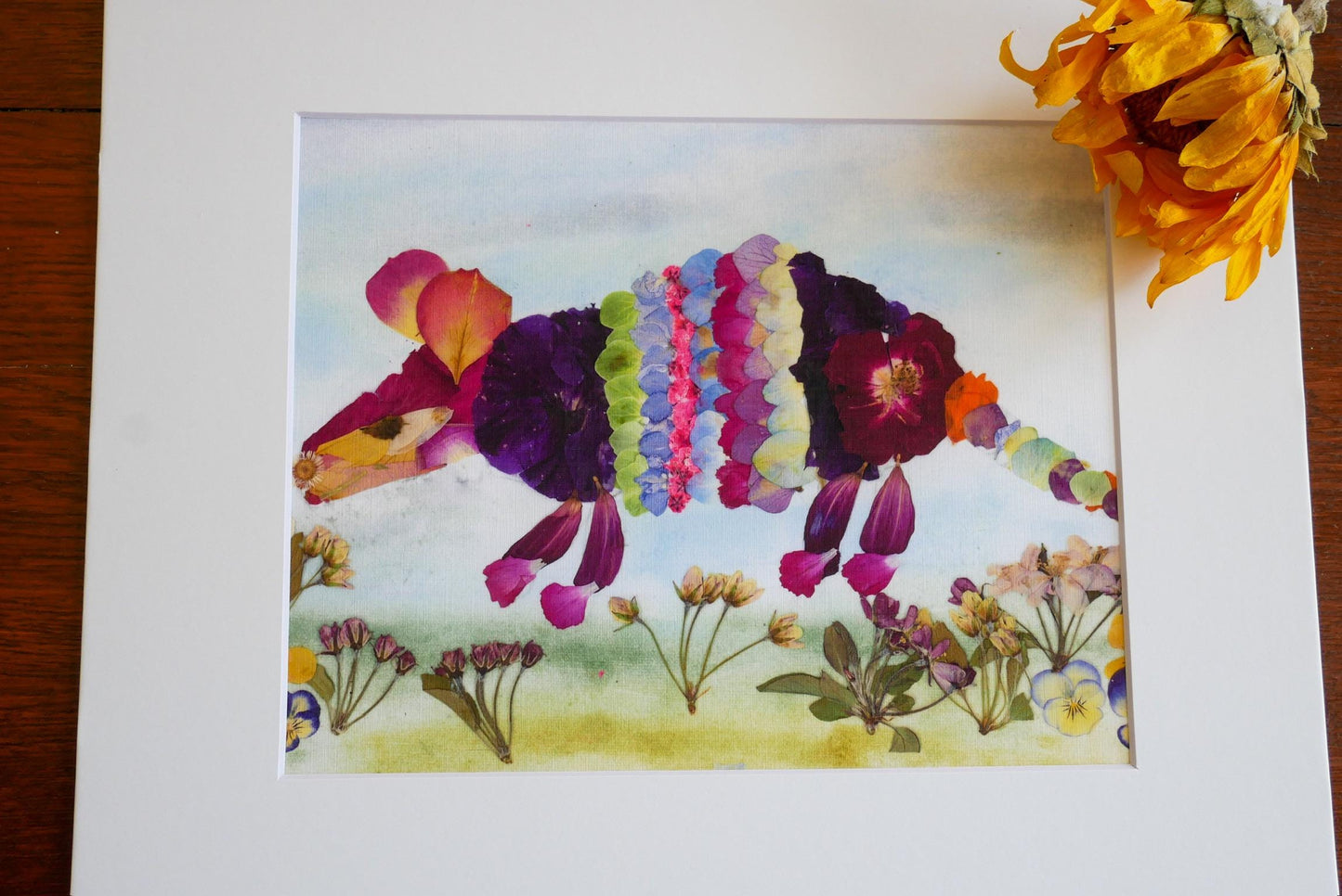 A framed artwork depicting a colorful, flower-like creature made from dried petals, surrounded by various flowers and plants.