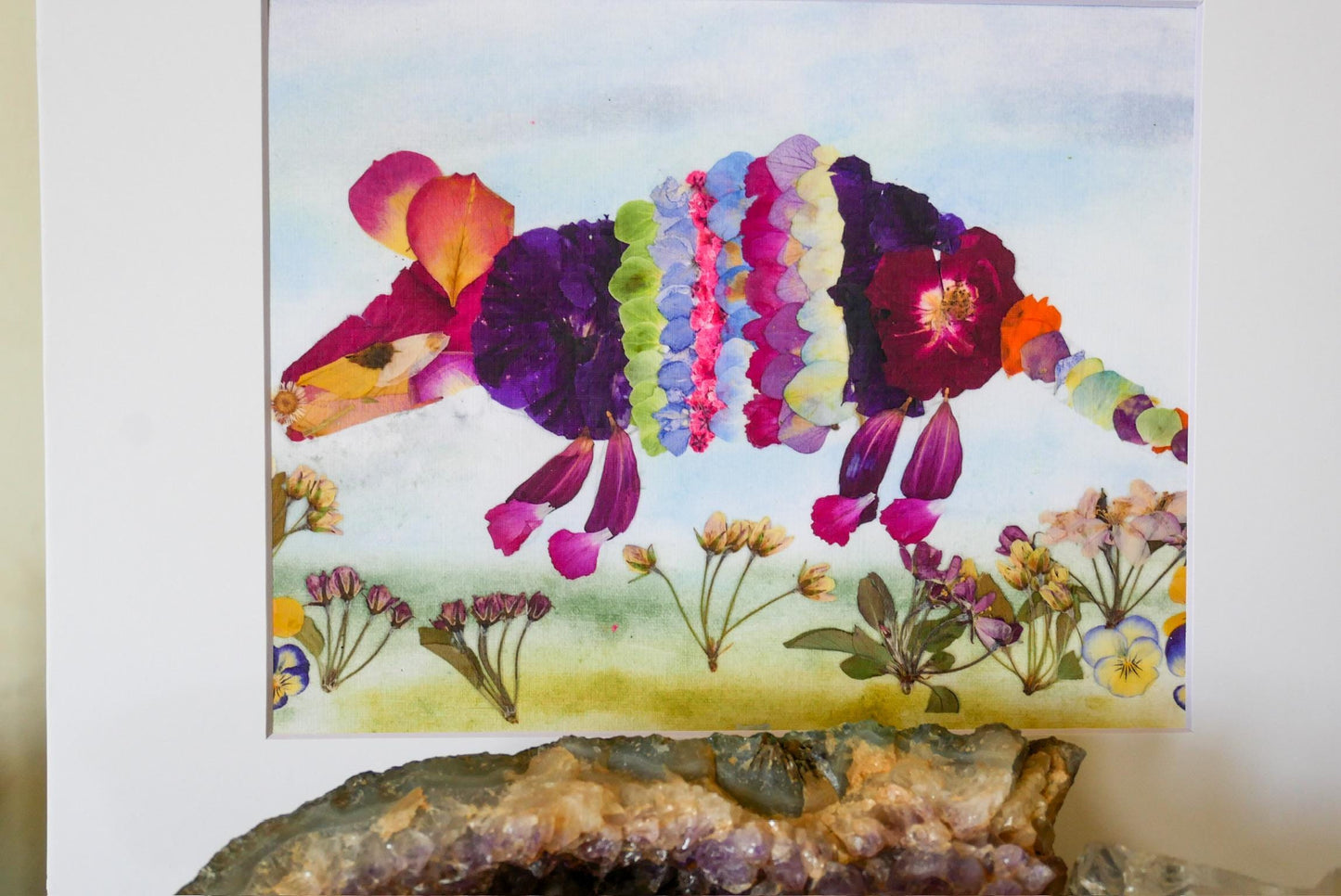 The image is a vibrant collage featuring a colorful, flower-like creature made from petals and leaves, set against a backdrop of various flowers and plants.