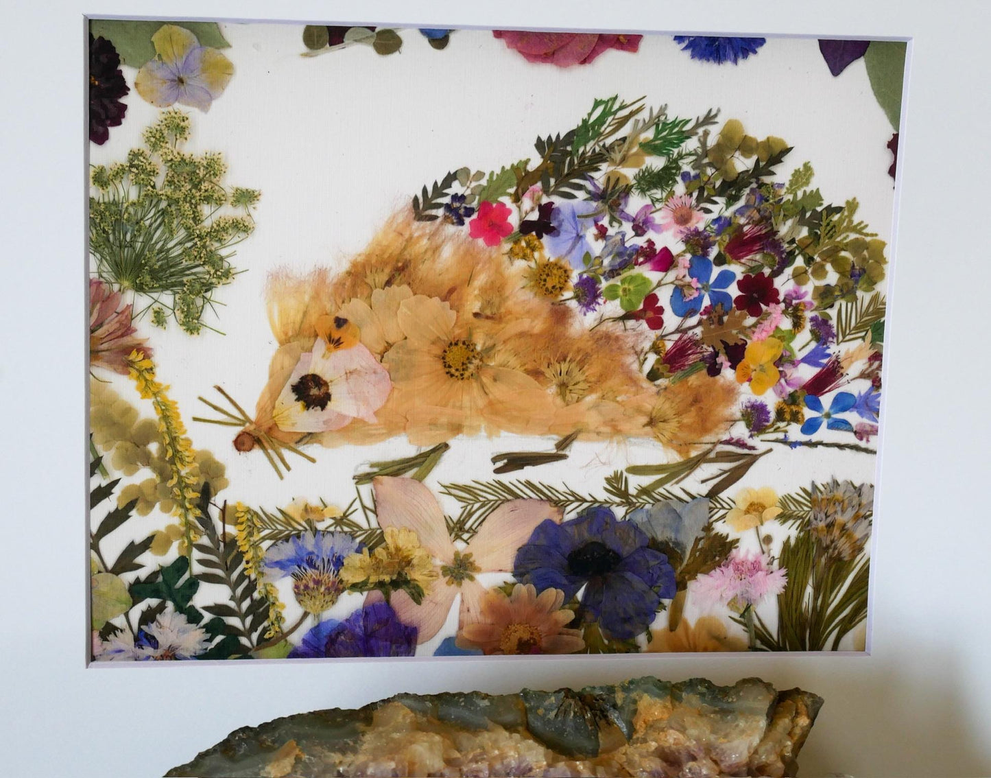 The image is a collage of various colorful flowers and plants, including a large orange flower, a variety of purple and pink flowers, and green leaves. The collage is framed and displayed on a wall, with a rock or stone visible in the foreground.