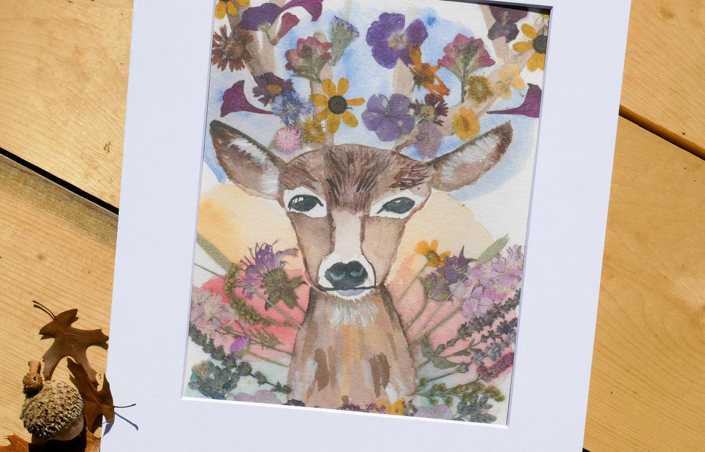 A framed watercolor painting depicting a deer with a floral crown on its head, surrounded by a vibrant array of flowers.