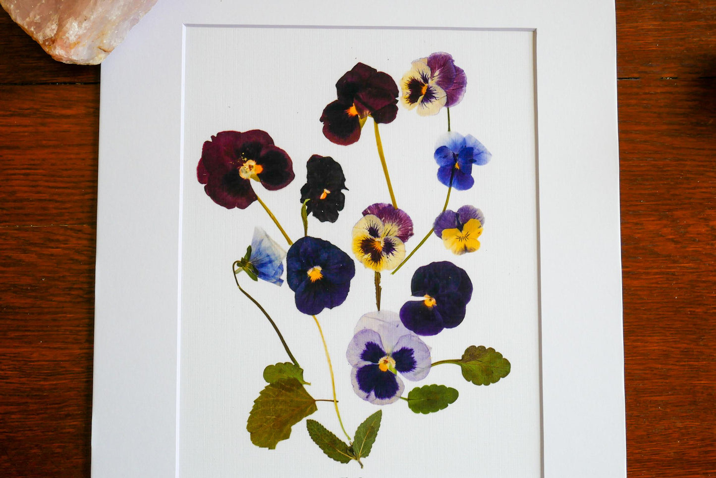 a white frame with a vibrant arrangement of colorful flowers, including purple, blue, and yellow pansies, along with green leaves.
