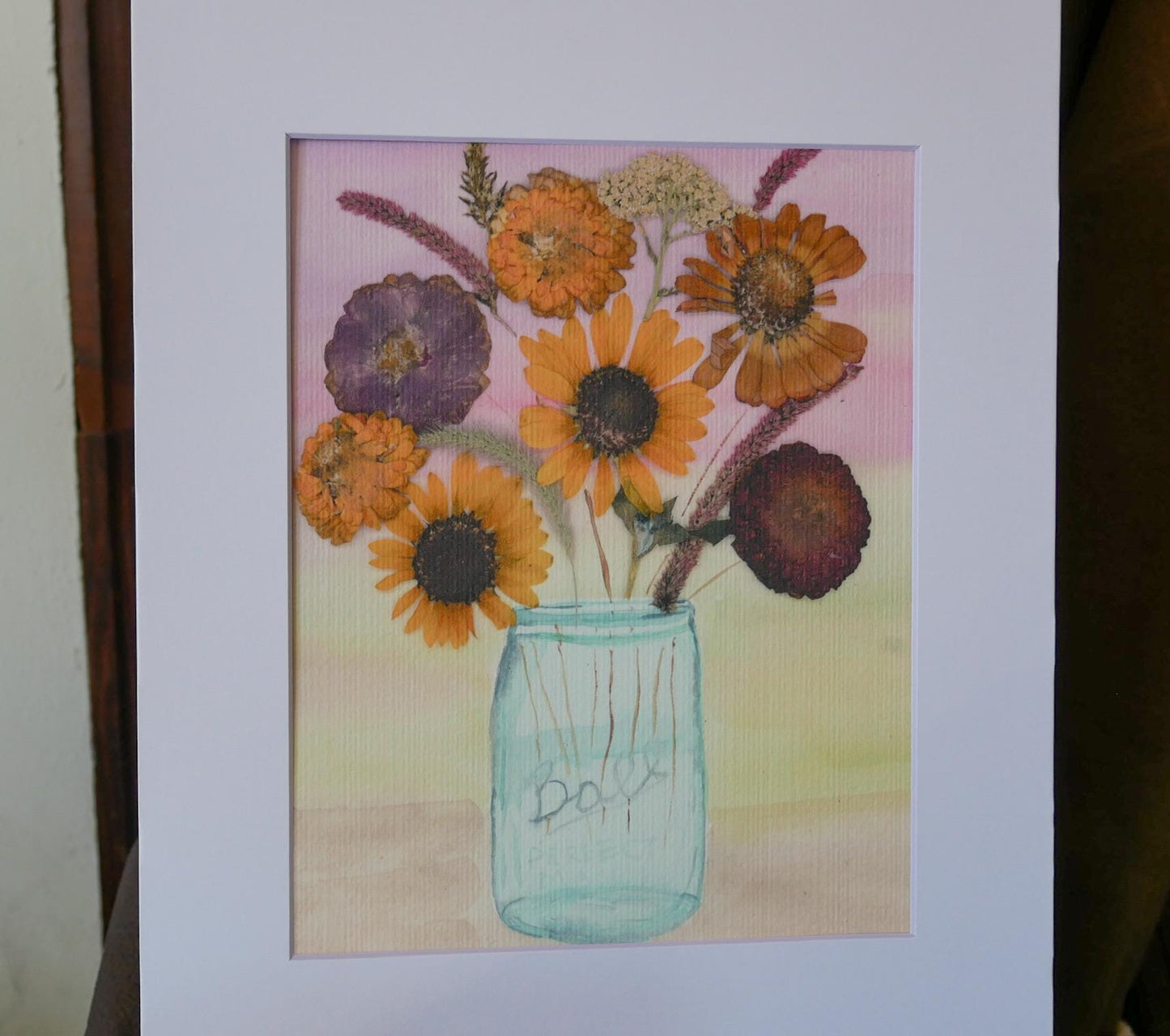 A framed painting depicting a vase filled with a variety of colorful flowers, including sunflowers and other blooms, set against a pink and yellow background.