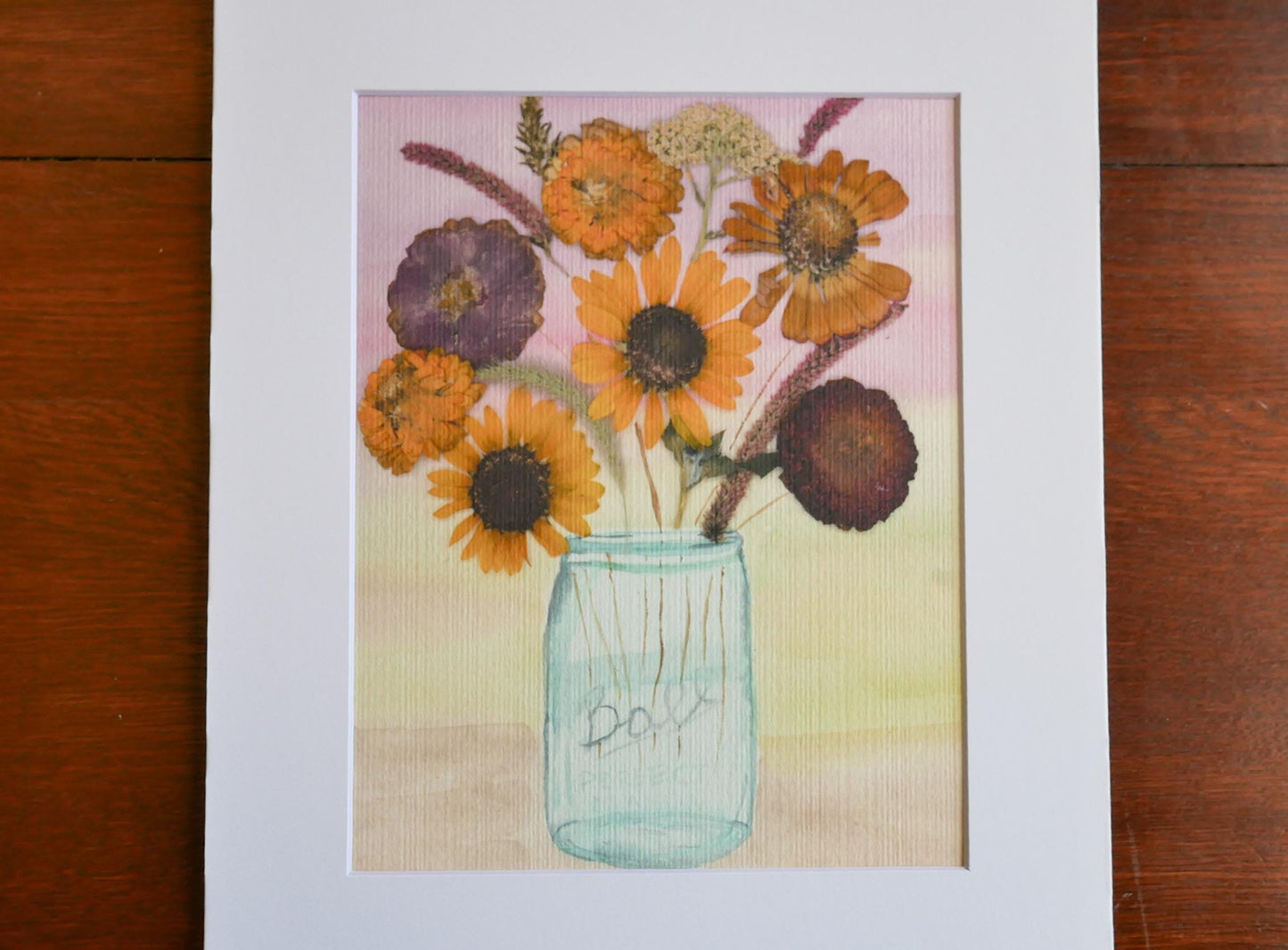 A framed watercolor painting depicting a vase filled with a variety of colorful flowers, including sunflowers and other blooms, set against a pink and yellow background.