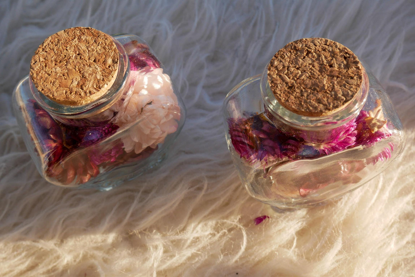 Two glass bottles with cork stoppers filled with pink and purple flowers, placed on a white furry surface.