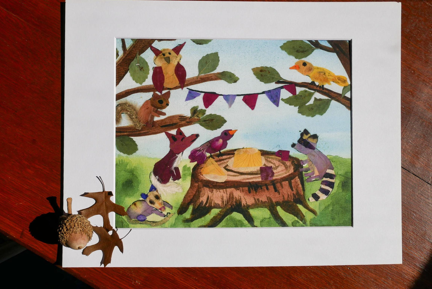 The image is a vibrant painting depicting a lively scene of various animals gathered around a tree stump, with a bird perched on a branch. The painting is framed and placed on a wooden surface, creating a warm and inviting atmosphere.