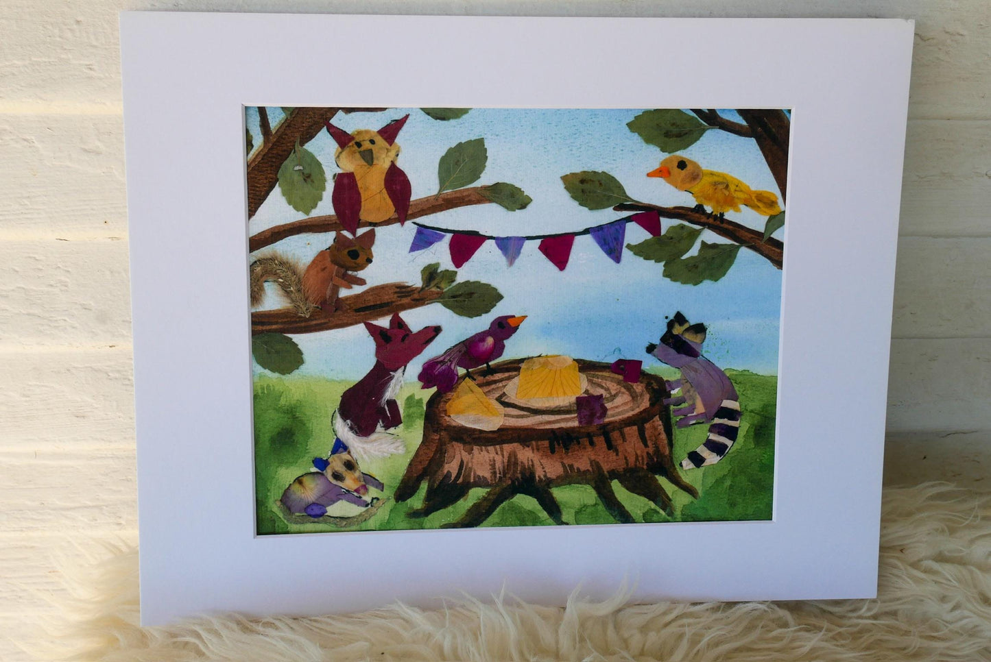 The image is a vibrant painting featuring a variety of colorful animals, including birds, squirrels, and a raccoon, gathered around a tree stump. The painting is framed and displayed on a white surface, with a fluffy white fur rug underneath.
