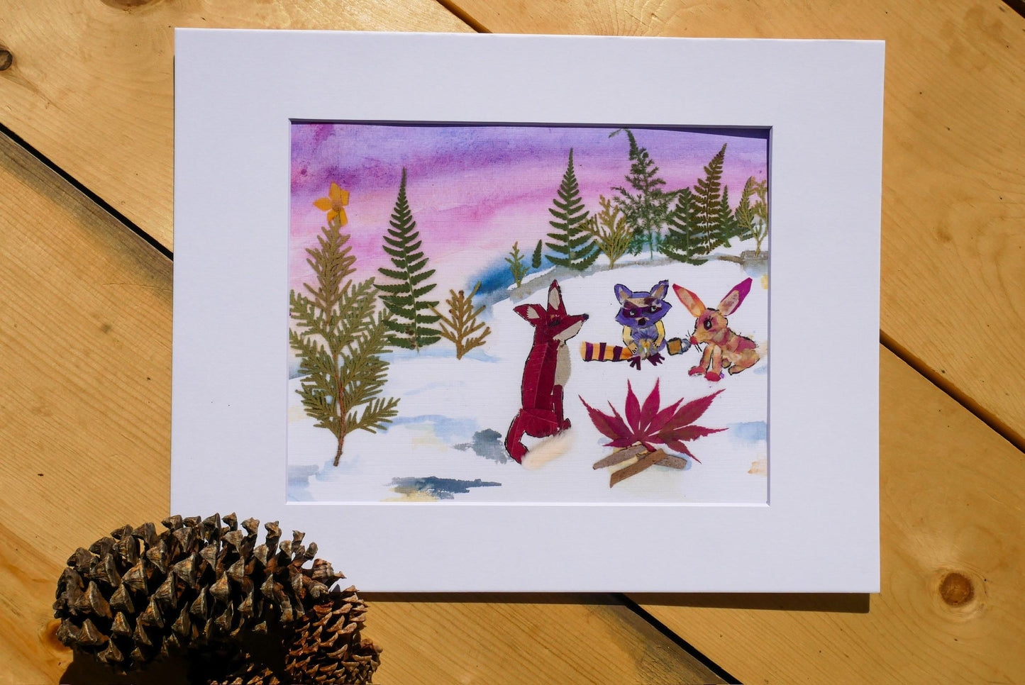 A framed watercolor painting depicting a snowy forest scene with animals, including a fox, rabbit, and deer, and a pine cone on a wooden surface.