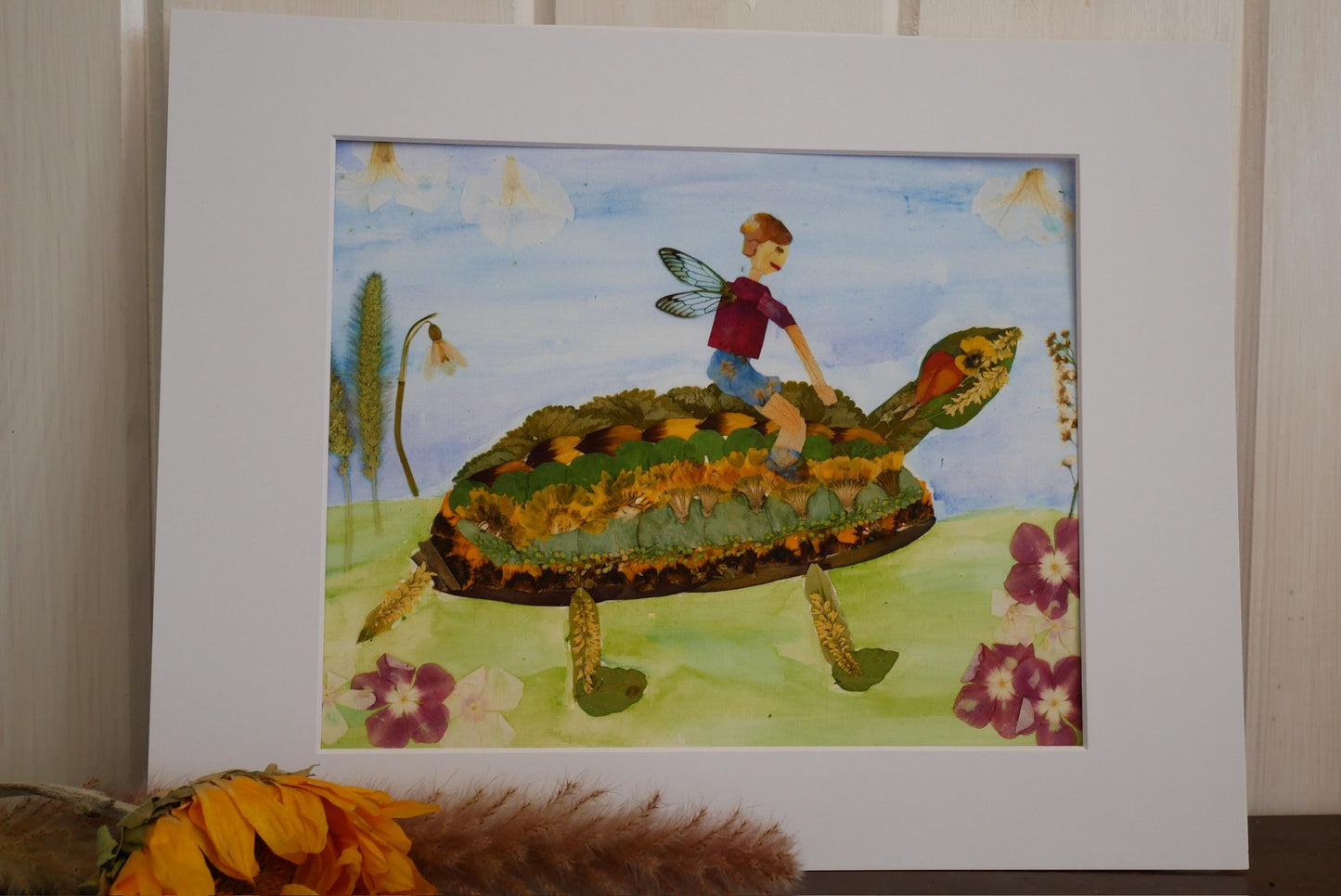 A framed artwork depicting a fairy riding a turtle through a garden with flowers and plants.