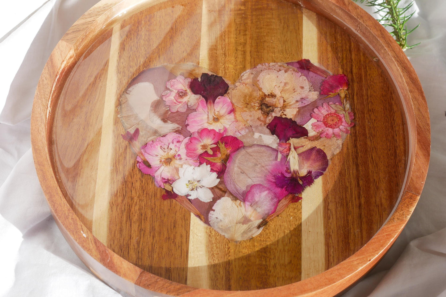 A wooden tray with a heart-shaped arrangement of dried pink and white flowers, including daisies and roses, placed on a white cloth.