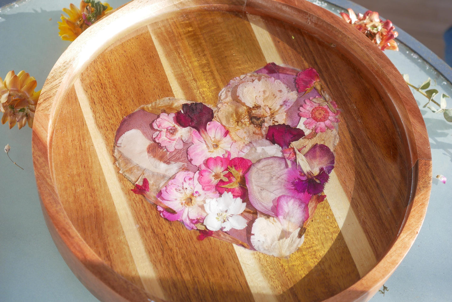 A wooden tray with a heart-shaped arrangement of dried pink and white flowers, surrounded by additional flowers and a blue surface.