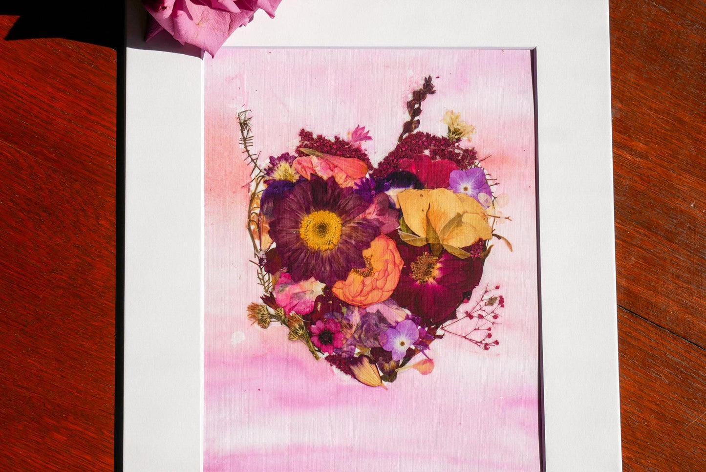 A framed artwork depicting a heart-shaped arrangement of various colorful flowers, including a large purple flower and smaller yellow and pink flowers.